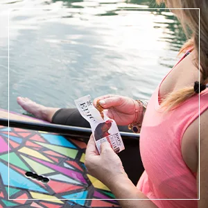 Epic Bacon Bits Hickory Smoked, 3 Ounce - Woman eating an EPIC grass fed protein bar while sitting on a paddle board