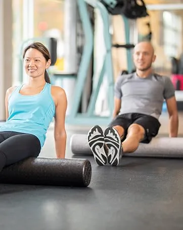 OPTP PRO Shoulder Support Shoulder Pillow for Rotator Cuff Pain Use During Recovery Following Surgery and Post Shoulder Sling - Post-Athletic Recovery Tool - Large/Extra Large - woman and man exercising on a black and silver foam roller in a gym setting