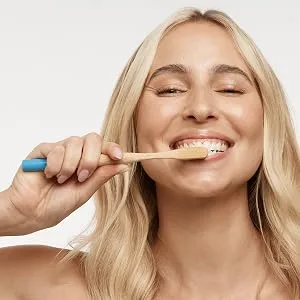 MooGoo Moothpaste - Natural Whitening Toothpaste with Hydroxyapatite for Adults & Kids Fluoride-Free Non-Toxic SLS-Free Vegan. Remineralizes & Removes Plaque - A blond woman smiling while brushing her teeth with a bamboo toothbrush