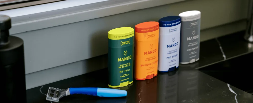 Mando Bourbon Leather Body Deodorant - 72 Hour Odor Control Aluminum Free Baking Soda Free - 3.0 Oz Tube and 2.6 Oz Solid Stick - MANDO Smooth Solid Deodorants on a bathroom countertop next to a shaver
