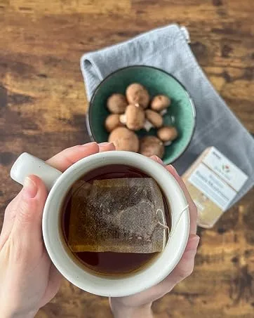 NOW Caring Tea Red Raspberry Tea Premium Unbleached Tea Bags with our no-staples design 24 Count - NOW Caring tea in a mug held by hands above a dining table with mushrooms