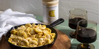 Aged White Cheddar Alfredo Pasta Sauce - 1 Jar - A cast iron pot with tortellini, aged white cheddar pasta sauce, and two glasses of red wine