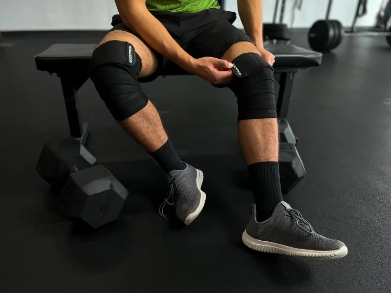 Bodyprox Knee Wrap 2 Pack for Squats, Weightlifting, Powerlifting, Leg Press, and Cross Training, Knee Support for Men and Women - Man sitting on bench wrapping his knees before working out