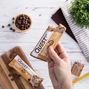 Quest Nutrition Birthday Cake Protein Bars, High Protein, Low Carb, Gluten Free, Keto Friendly, 12 Count - Image of hand holing cookie dough bar with chocolate chips in the background