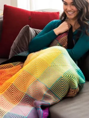 Knit Picks Hue Shift Afghan Knitting Pattern Kit (Rainbow) - A model snuggled up under the Hue Shift blanket