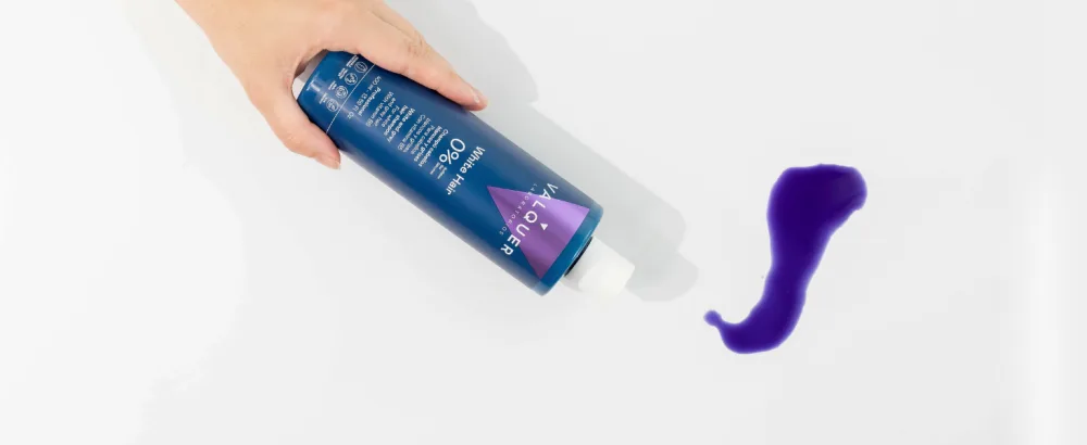 Valquer Laboratorios V lquer Shampoo for white and gray hair 1000 ml - Hand met een blauwe spuitfles naast een paars, gebogen voorwerp op een lichte achtergrond.