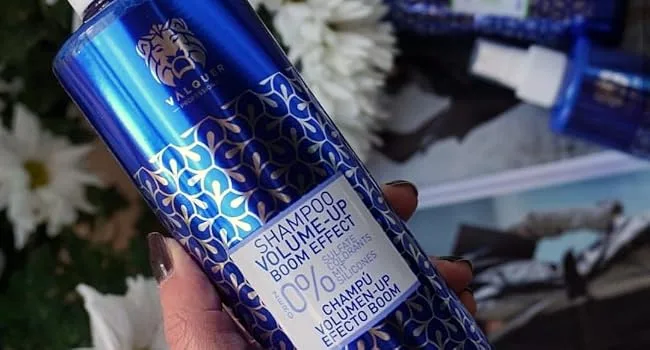 Valquer Laboratorios V lquer Shampoo VolumeUp Boom Effect 400 ml - Metaalblauwe fles met geometrisch patroonontwerp met product voor persoonlijke verzorging, afgebeeld tegen witte bloemen op de achtergrond.
