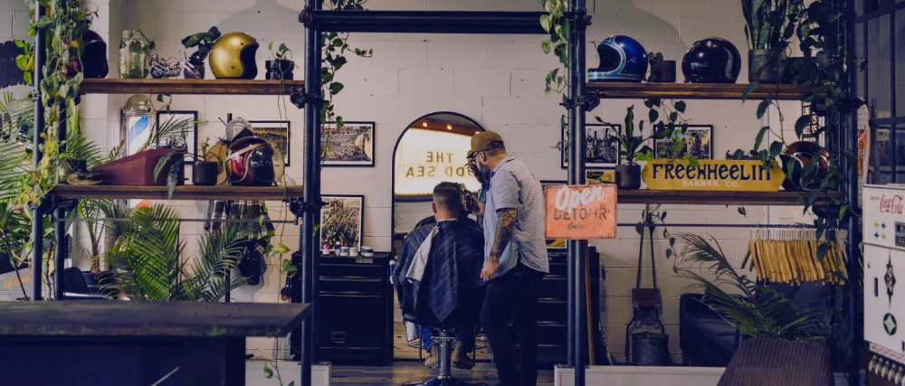 Shear Revival Younger Day Pomade 4 oz - Interieur van een kapperszaak met een inrichting in vintage-stijl. Op houten planken staan planten en kappersgereedschap. Een klant zit in een stoel terwijl een kapper werkt, omringd door retro signalisatie en apparatuur