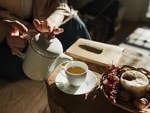 Your ExcellenTEA Earl Grey Blue Loose Leaf Black Tea Bergamot Ceylon Black Tea with Cornflower Petals Floral Earl Grey Blend - 250g  - White ceramic teapot pouring tea into a cup on saucer. Wooden table with bread and red berries visible. Cozy, warm-toned setting.