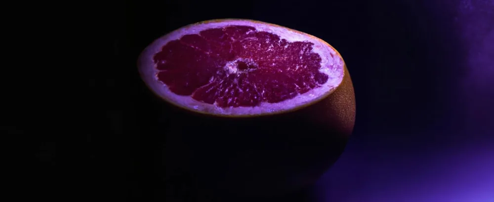 Naturally Wicked Grapefruit Lip Scrub | Natural & Vegan Sugar Exfoliant For Lips | 15ml - Fresh Chopped Pink Grapefruit With Red Flesh Exposed Under Mysterious Purple Light